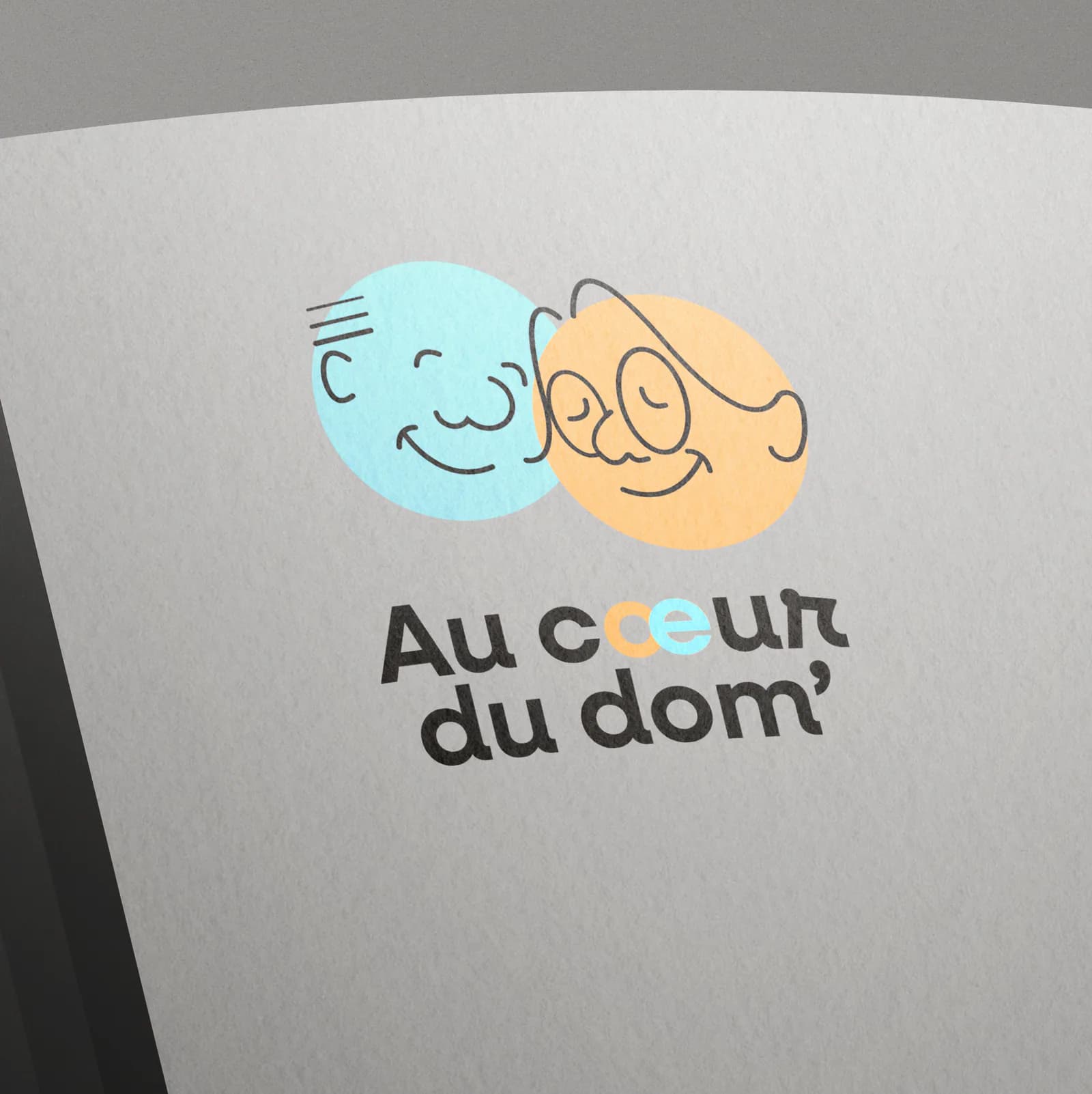 Logo Au cœur du dom’ imprimé sur une pile de feuilles gris clair : deux visages souriants dans des cercles bleu et orange, au-dessus du nom.