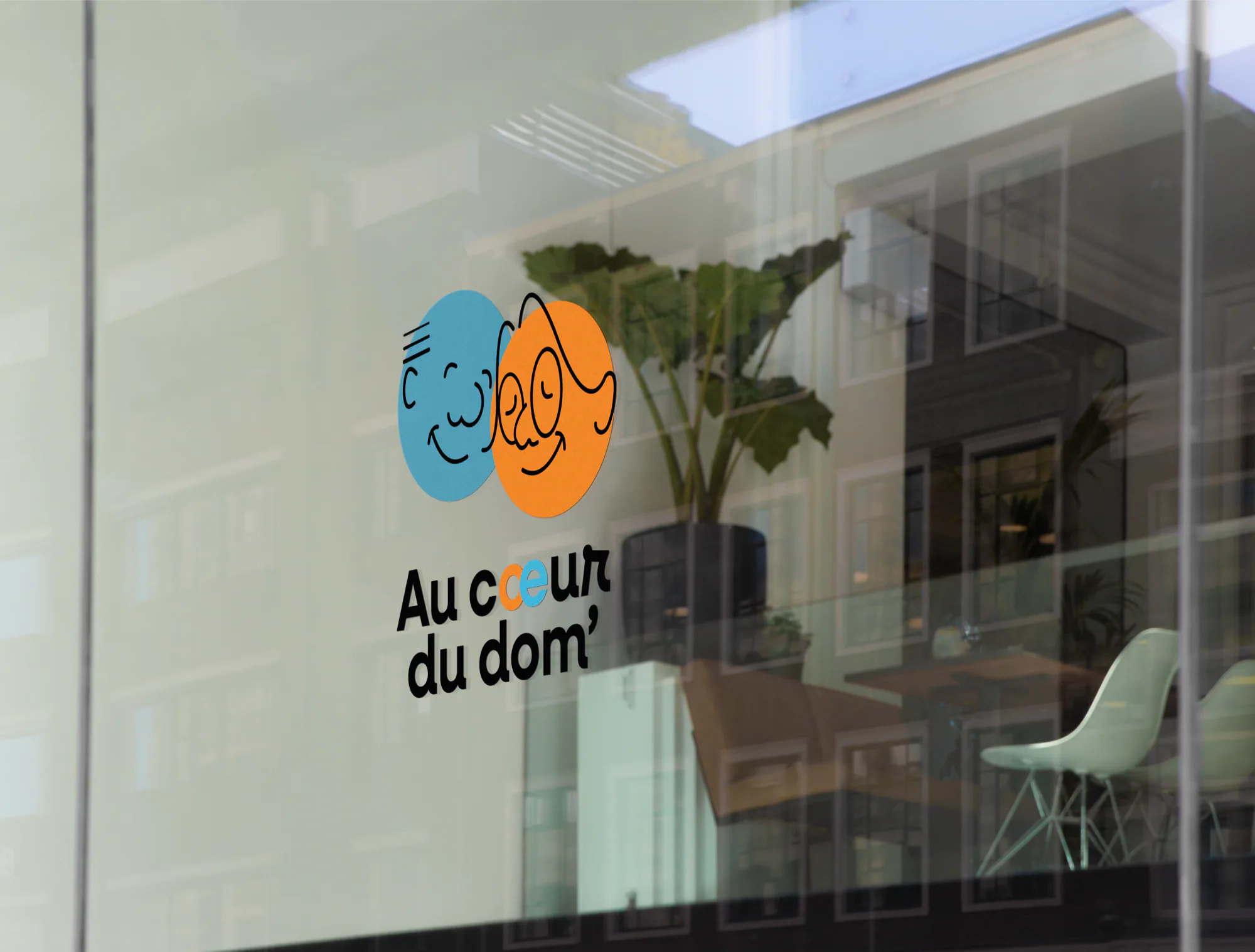 Logo Au cœur du dom’ collé sur une vitrine : deux visages dans des cercles bleu et orange, avec un bureau et une grande plante verte derrière la vitre.