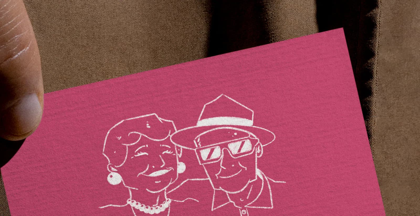 Détail du recto rose vibrant tenu en main : portraits d’Amélie et Robert tracés en blanc sur un papier texturé, devant un tissu brun