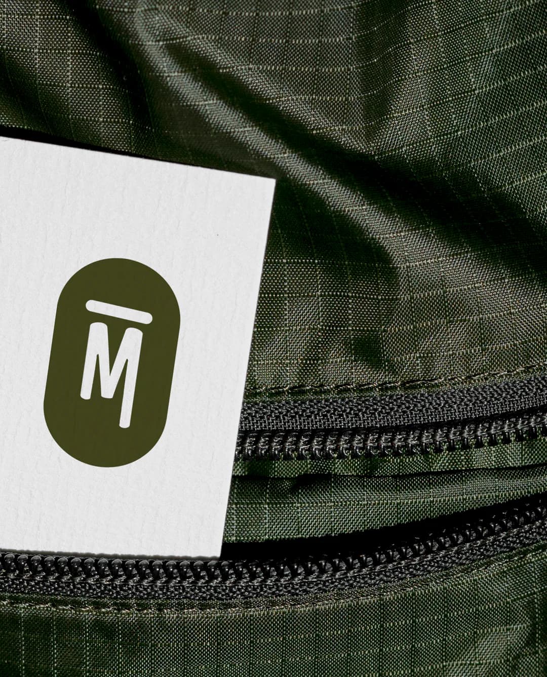 Gros plan sur une carte de visite blanche glissée dans la poche zippée d’un sac en nylon vert, avec le monogramme La PAMPA en vert olive
